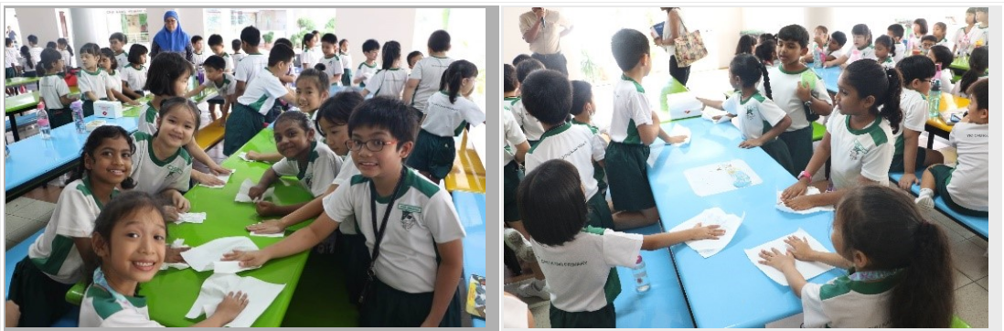 Young students in white and green uniforms sit at colorful tables, handling papers during a school activity.